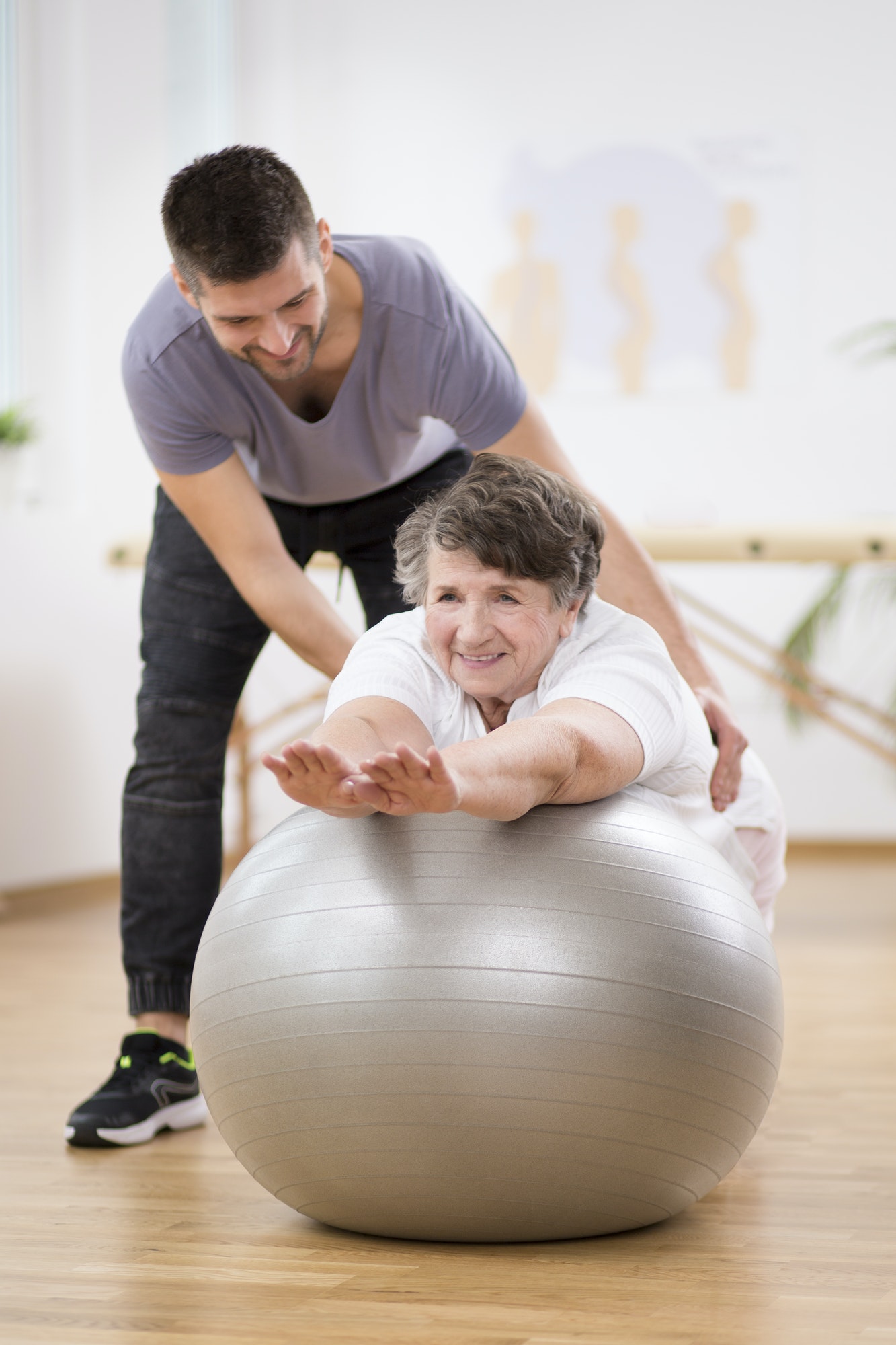 Smiling physiotherapy student helping senior woman lay on the exercising ball during rehabilitation